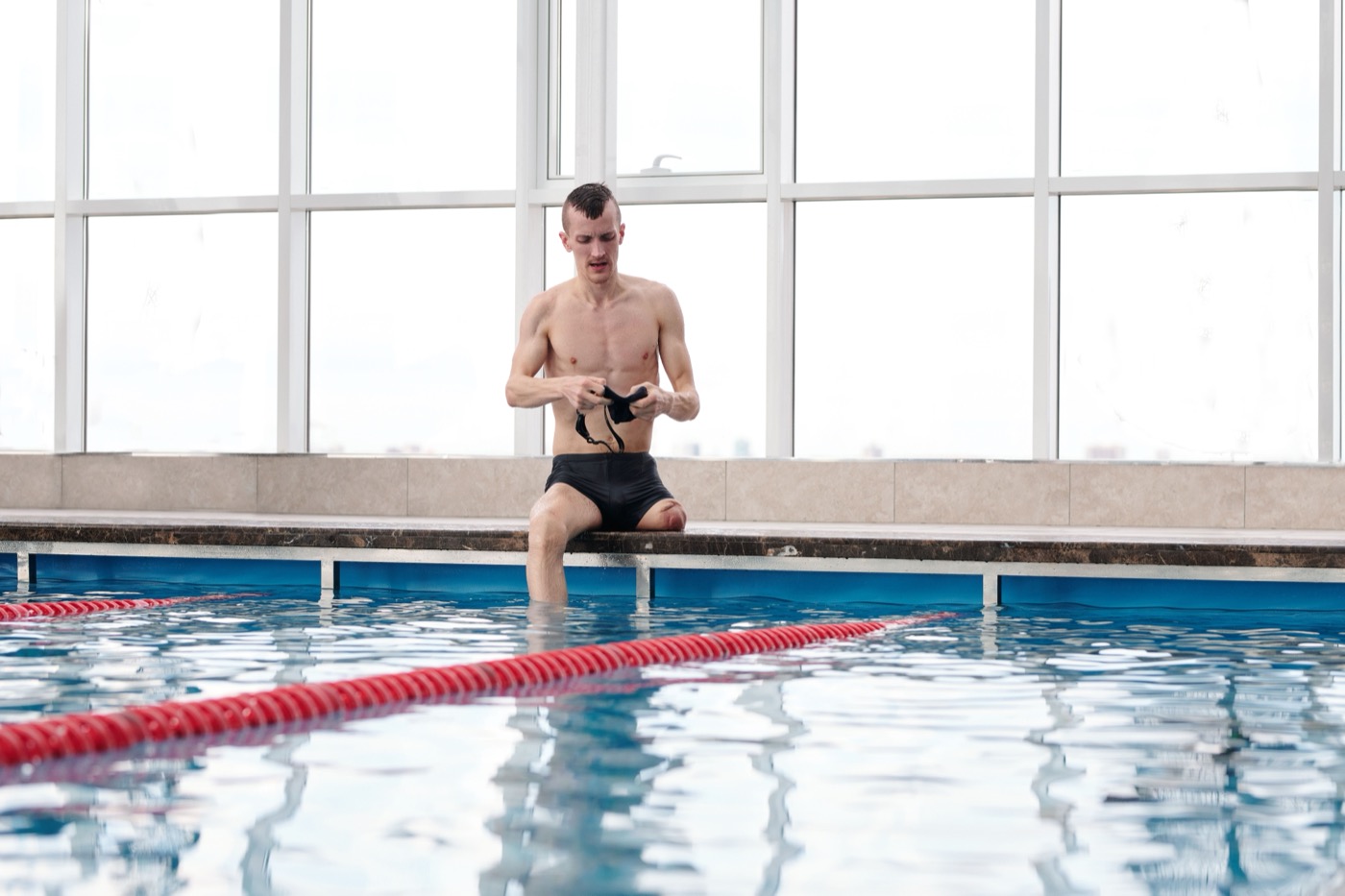 A swimmer with a lower-leg limb difference sitting at the pool edge adjusting his goggles before a session
